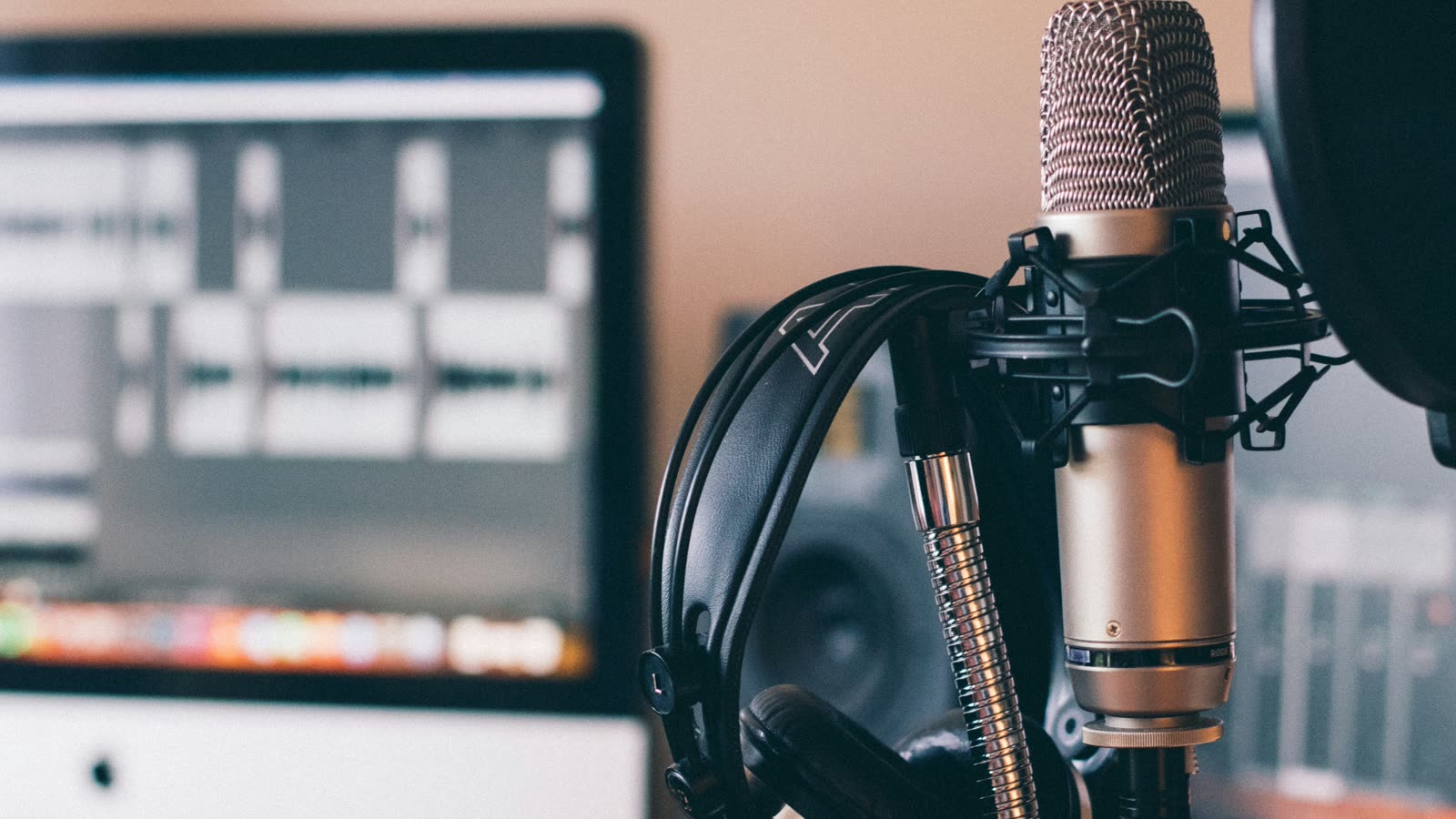 A studio microphone with headphones hanging on its stand in front of a monitor showing an audio editor