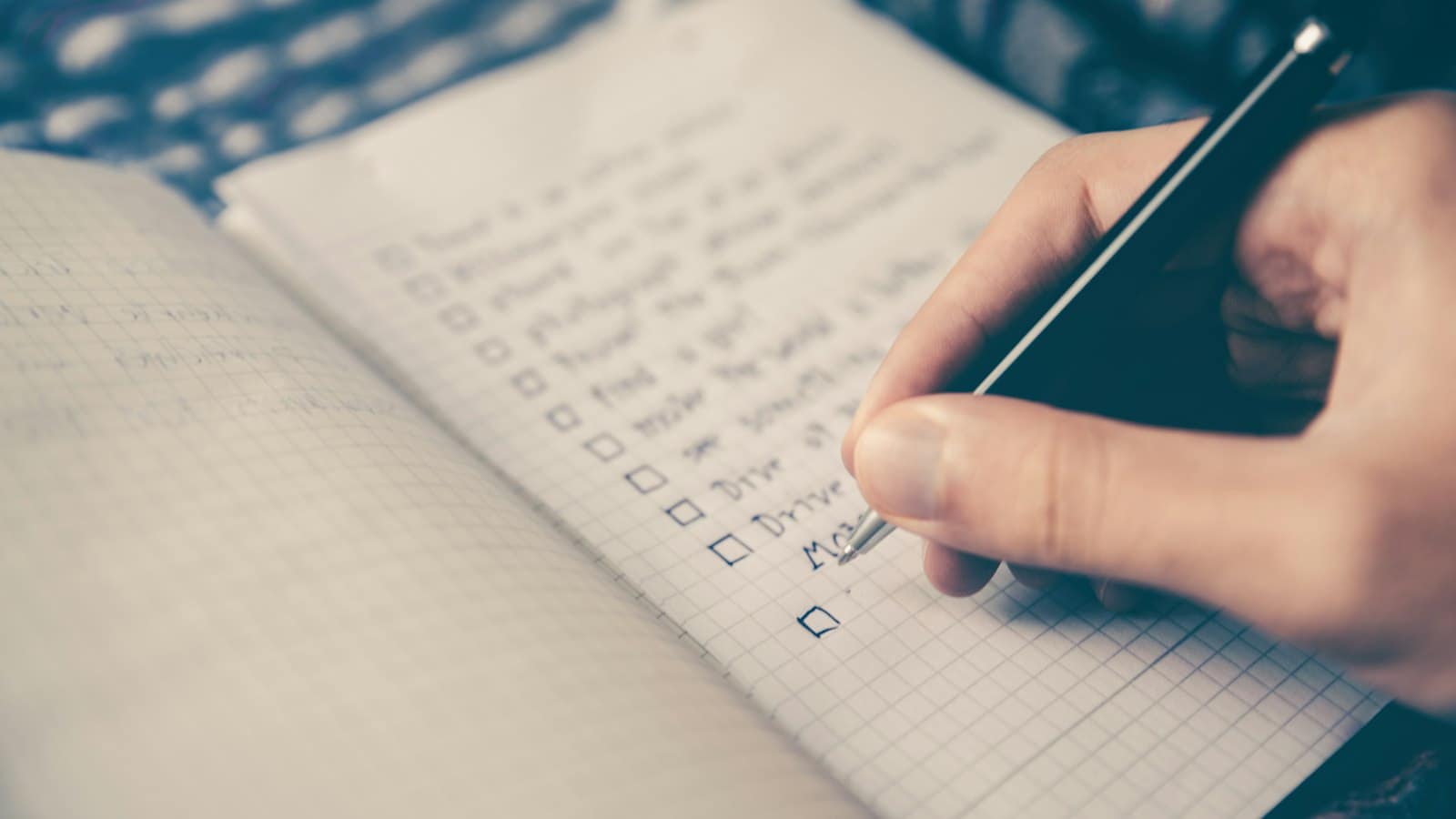 A hand with a pen writing a checklist in a squared notebook