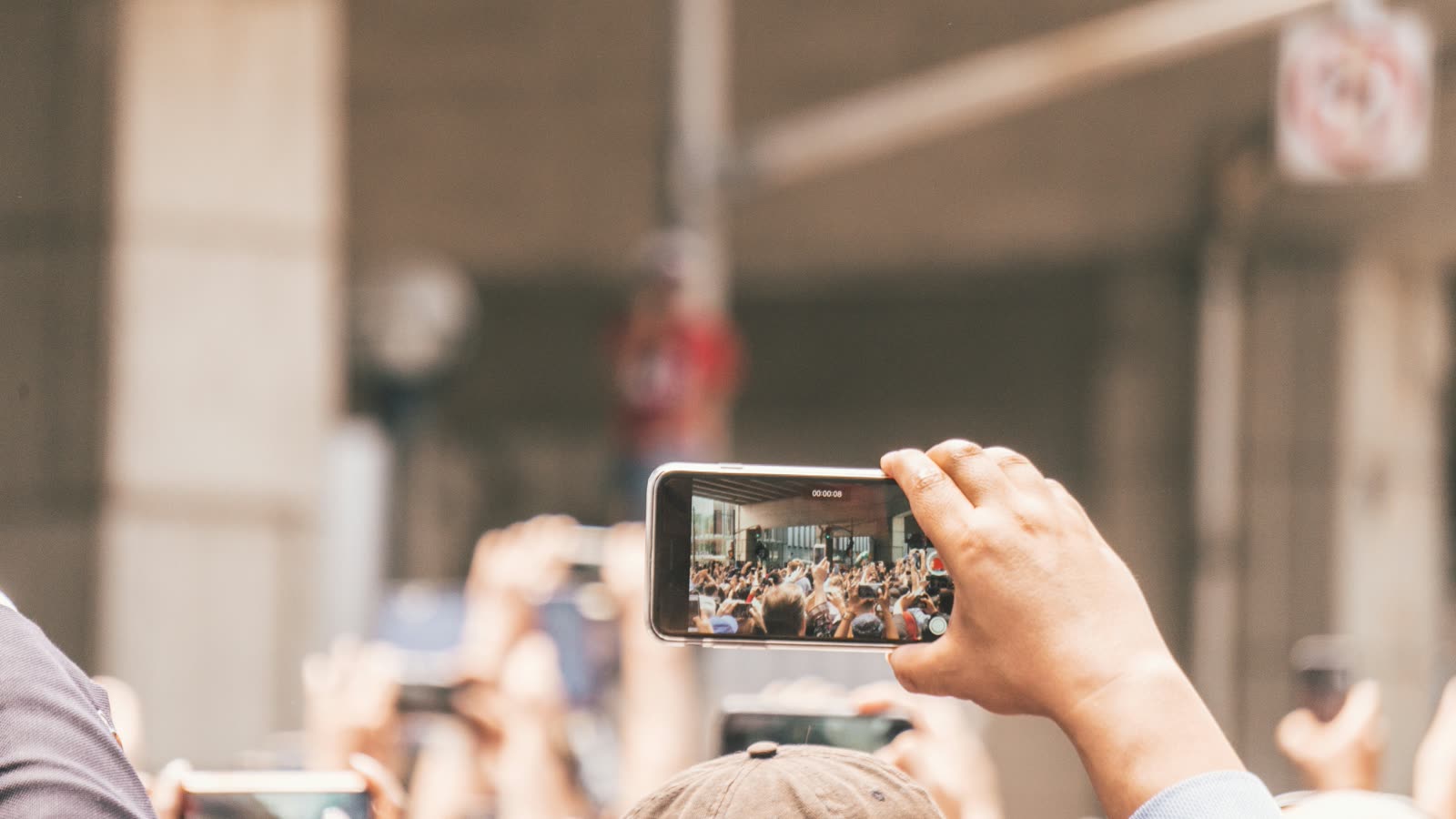 A hand holding up a phone to film a crowd, the crowd showing on the phone screen