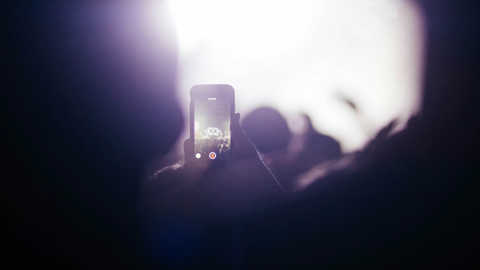 A hand holding up a phone recording a video in a dark, crowded room